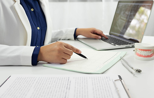 The image depicts a person in a medical professional s attire, seated at a desk with a laptop, writing on paper, and surrounded by office supplies.