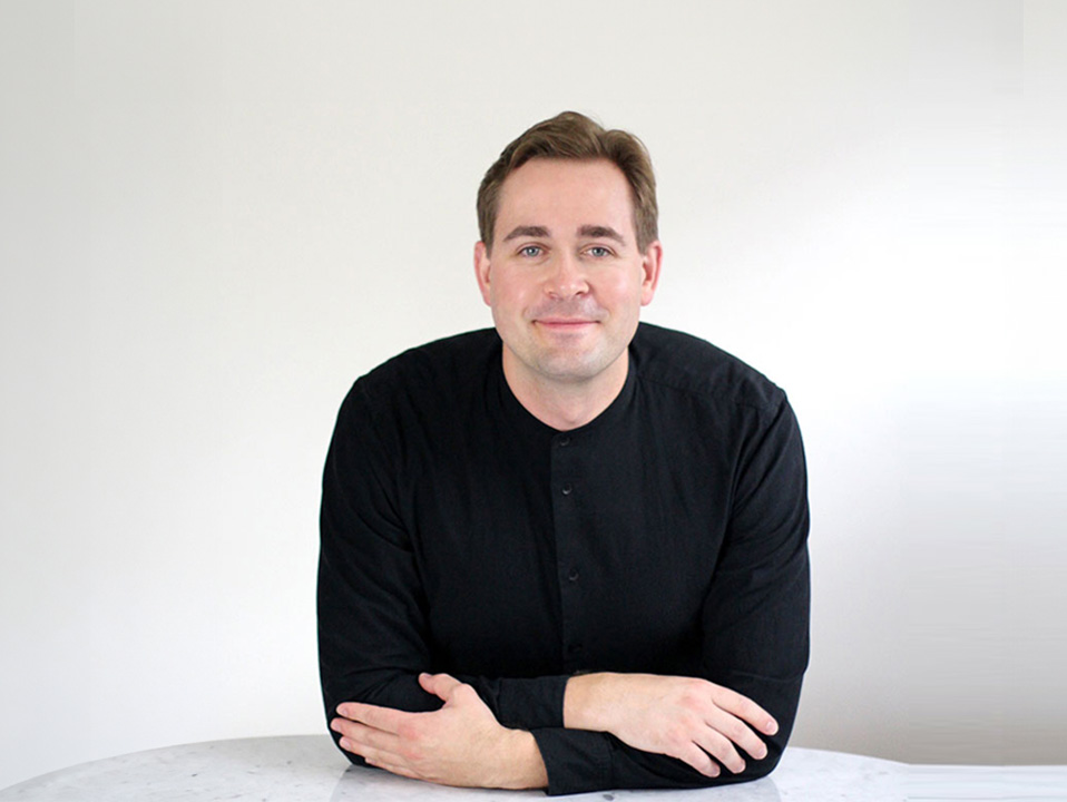 The image shows a man sitting at a table, leaning slightly forward with his arms crossed. He is wearing a black shirt and has short hair. The background is plain and light-colored, providing a neutral backdrop that contrasts with the subject.